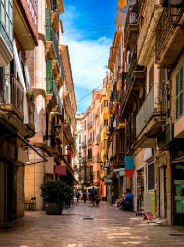 Few tourists walk through a narrow mediterranean alley with small shops and restaurants in the shadow of residential buildings in the groomed centre of Palma de Mallorca on an idyllic day in summer.